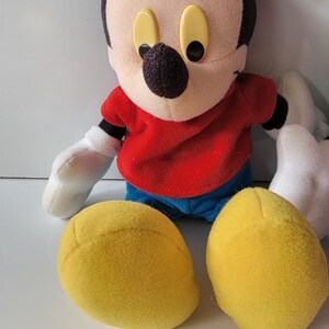 May include: A plush Mickey Mouse toy with black ears, a red shirt, blue jeans, and yellow shoes. The toy has a black nose and large yellow eyes.
