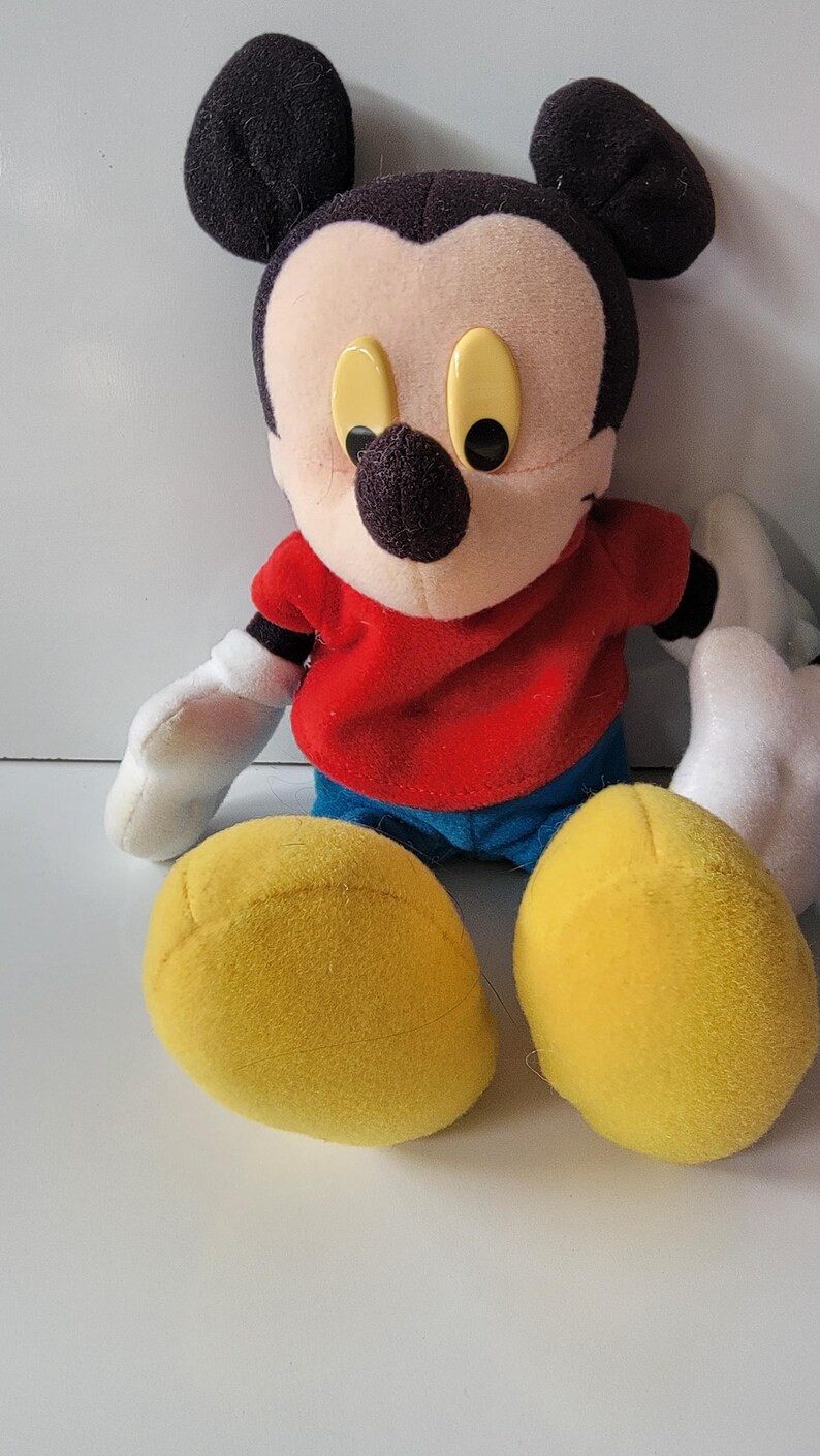 May include: A plush Mickey Mouse toy with black ears, a red shirt, blue jeans, and yellow shoes. The toy has a black nose and large yellow eyes.