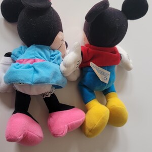 May include: A pair of plush Mickey Mouse and Minnie Mouse toys. Mickey is wearing a red shirt and blue trousers. Minnie is wearing a blue dress with a pink bow.