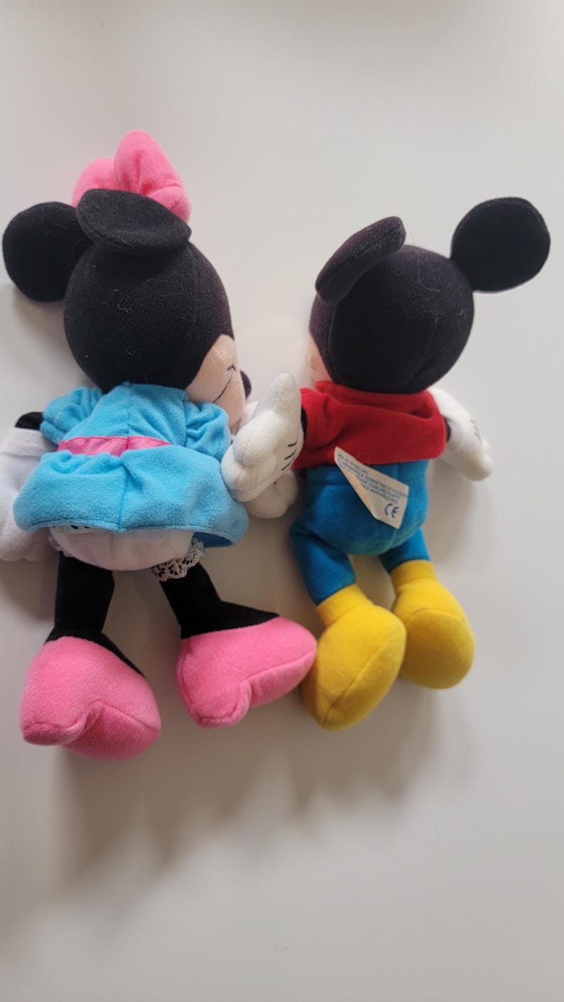May include: A pair of plush Mickey Mouse and Minnie Mouse toys. Mickey is wearing a red shirt and blue trousers. Minnie is wearing a blue dress with a pink bow.