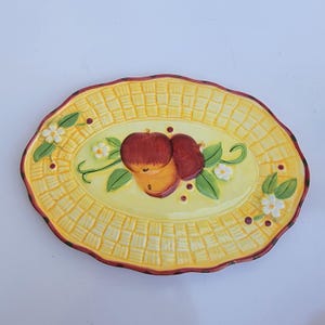 May include: Oval-shaped ceramic serving platter with a yellow basket weave design. The center features a painted illustration of red and yellow apples with green leaves and white flowers. The rim is a dark red color.
