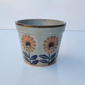 May include: A ceramic planter with a speckled light gray glaze and a dark brown rim. The exterior features a repeating pattern of orange-toned sunflower motifs with dark centers, complemented by dark blue stems and leaves at the base.