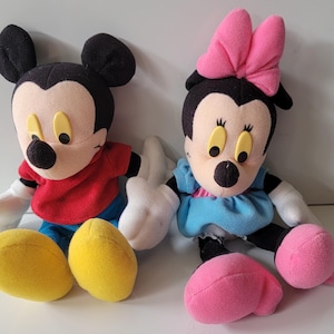 May include: A plush Mickey Mouse and Minnie Mouse toy set. Mickey is wearing a red shirt and blue trousers. Minnie is wearing a blue dress with a pink bow.