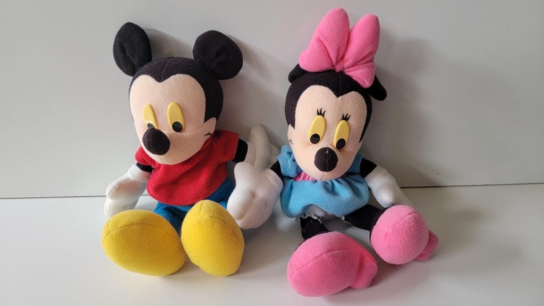 May include: A plush Mickey Mouse and Minnie Mouse toy set. Mickey is wearing a red shirt and blue trousers. Minnie is wearing a blue dress with a pink bow.