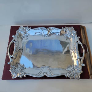 May include: A rectangular silver-toned serving tray with a polished, reflective surface. The tray features ornate, raised scrollwork and fleur-de-lis style motifs at each corner, along with integrated curved handles on the left and right sides.