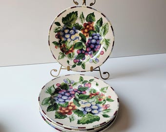 May include: A set of four ceramic plates with a grape design. The plates feature clusters of purple, blue, and red grapes with green leaves on a cream background. A decorative metal plate stand holds one plate.