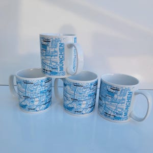 May include: Four white ceramic mugs with a blue map design of Munich. The mugs have a curved handle and the word "MUNCHEN" is printed vertically on the side. The map design includes street names and landmarks.