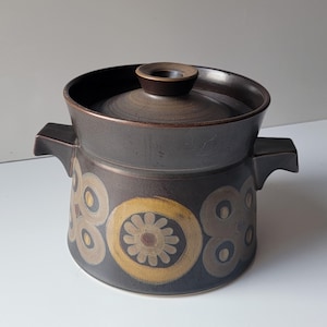 May include: A dark brown ceramic casserole dish with a lid. The dish features a floral pattern in shades of tan and gold. It has two handles and a lid with a small, round knob. The dish is likely vintage or retro.