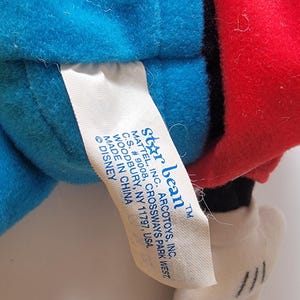 May include: A blue and red plush toy of Mickey Mouse with a white tag that reads "Star Bean&trade; Mattel, Inc. Arcotoys, Inc. C.S. # 9008, Crossways Park West, Woodbury, NY 11797, USA. Made in China &copy; Disney"