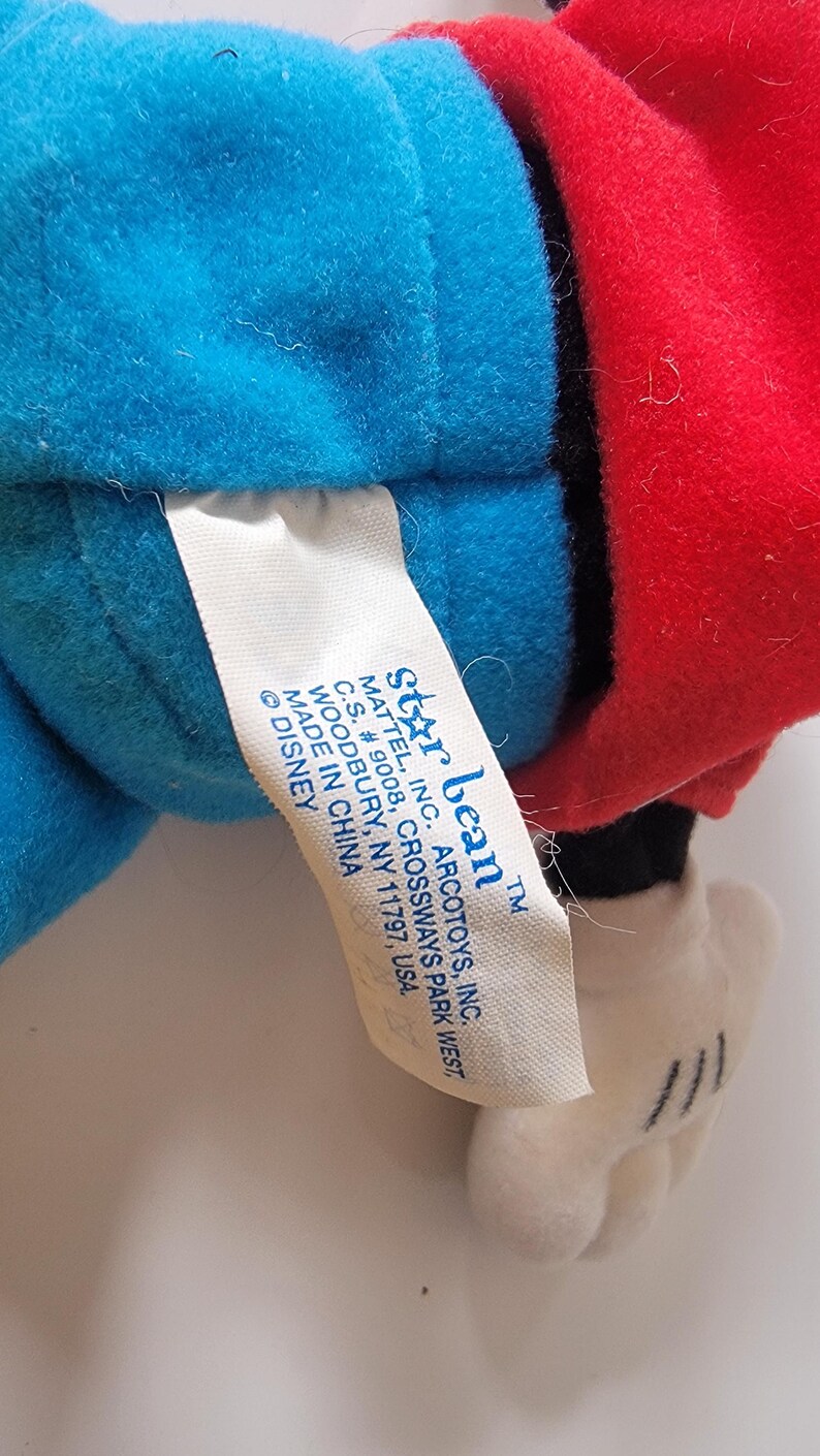 May include: A blue and red plush toy of Mickey Mouse with a white tag that reads "Star Bean&trade; Mattel, Inc. Arcotoys, Inc. C.S. # 9008, Crossways Park West, Woodbury, NY 11797, USA. Made in China &copy; Disney"