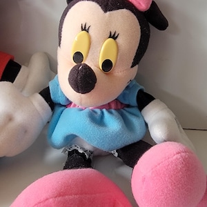 May include: A plush toy of Minnie Mouse wearing a blue dress with a pink bow on her head. She has black eyes and yellow pupils. Her shoes are pink.
