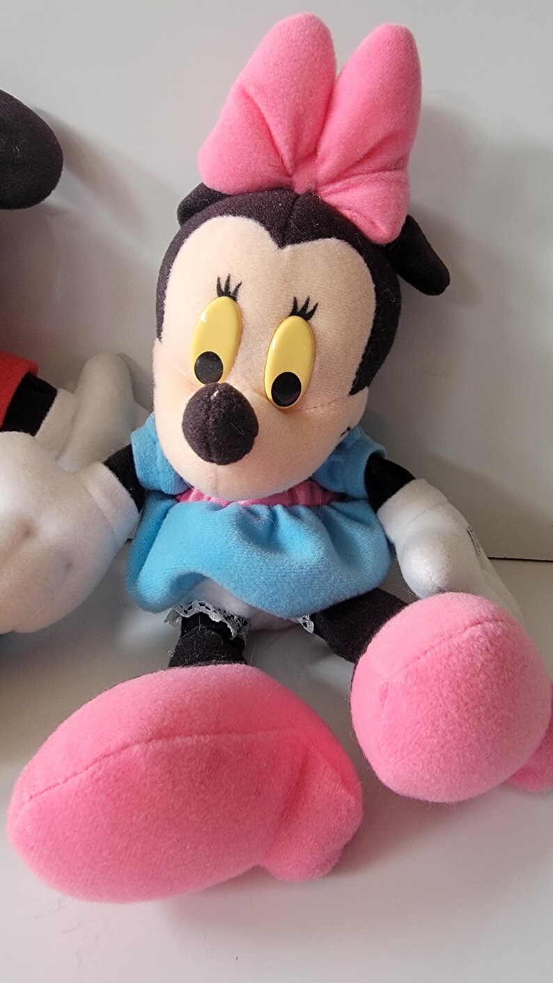 May include: A plush toy of Minnie Mouse wearing a blue dress with a pink bow on her head. She has black eyes and yellow pupils. Her shoes are pink.