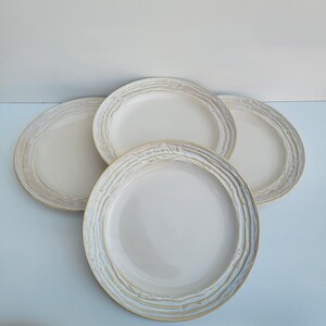 May include: Four off-white ceramic plates with a textured, ridged border. The plates are arranged in a circular pattern, showcasing their simple yet elegant design. The plates are suitable for serving meals.