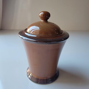 May include: A brown ceramic jar with a lid. The jar has a rounded base and a flared body, with a dark brown rim and lid. The lid has a small, round knob. The jar is likely used for storage.