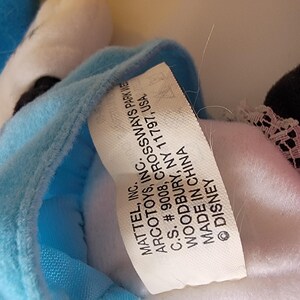 May include: A white tag with black text attached to a blue and white plush toy. The tag reads "MATTEL, INC. ARCOTOYS, INC. C.S. # 9008, CROSSWAYS PARK WEST WOODBURY, NY 11797, USA. MADE IN CHINA &copy; DISNEY"