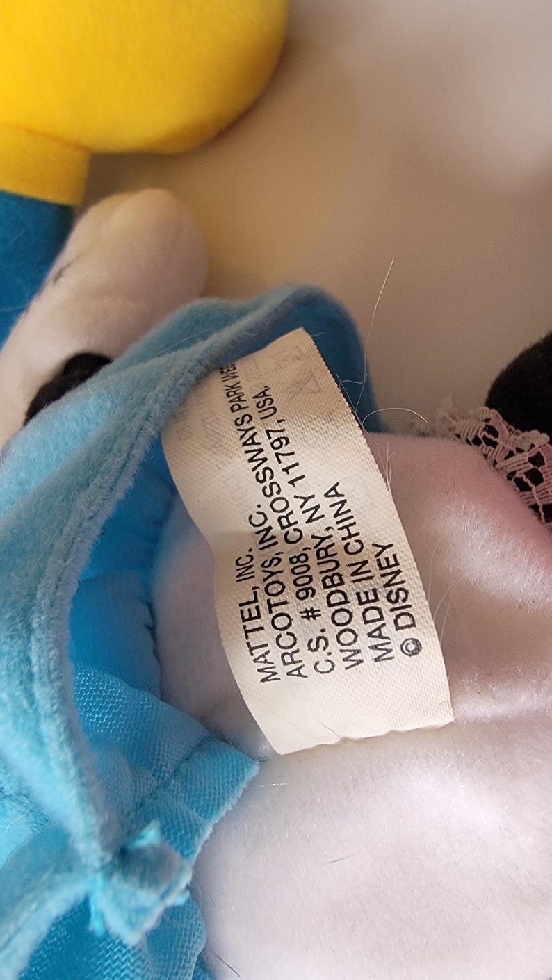 May include: A white tag with black text attached to a blue and white plush toy. The tag reads "MATTEL, INC. ARCOTOYS, INC. C.S. # 9008, CROSSWAYS PARK WEST WOODBURY, NY 11797, USA. MADE IN CHINA &copy; DISNEY"