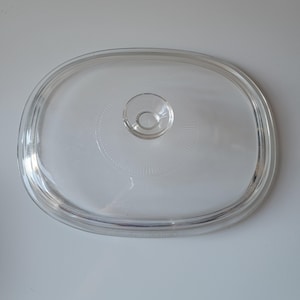 May include: Clear, oval-shaped glass lid with a small, round handle in the center. The lid has a raised rim and a textured design radiating from the handle. A kitchenware item.