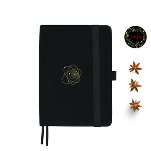 May include: A black notebook with a gold atom design and a black elastic closure. The notebook has a black elastic band and a black ribbon bookmark.