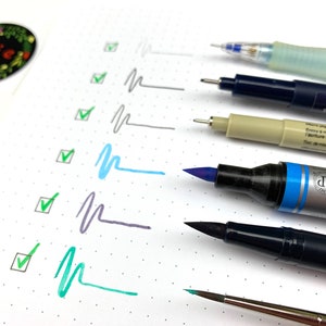 May include: A close-up of a variety of pens and markers, including a blue brush pen, a black brush pen, a grey marker, a beige marker, a blue and clear pen, and a silver and blue marker. The pens are arranged vertically on a white surface with a dotted pattern. The pens are all different sizes and have different nibs.