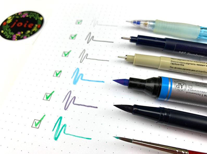 May include: A close-up of a variety of pens and markers, including a blue brush pen, a black brush pen, a gray marker, a beige marker, a blue and clear pen, and a silver and blue marker. The pens are arranged vertically on a white surface with a dotted pattern. The pens are all different sizes and have different tips.
