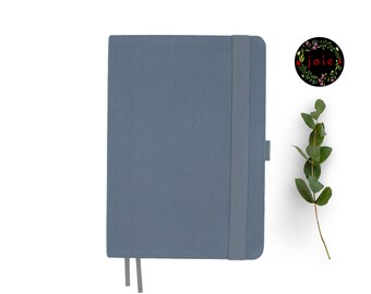 Puede incluir: Un cuaderno azul con cierre elástico gris y un marcador de cinta gris. El cuaderno está sobre un fondo blanco.