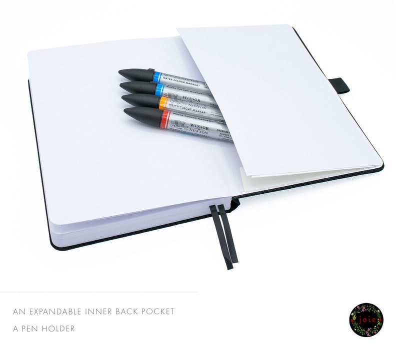 May include: A black notebook with an expandable inner back pocket and a pen holder. The notebook is open and has a white interior. There are four markers in the pocket, including a blue, orange, and two gray markers. The markers have the text "Winsor Newton Water Colour Markers" printed on them.