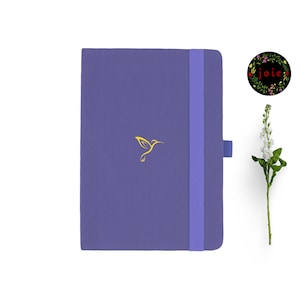 May include: A purple notebook with a gold hummingbird design and a purple elastic closure. The notebook has the word "joie" written in a floral wreath on the cover.