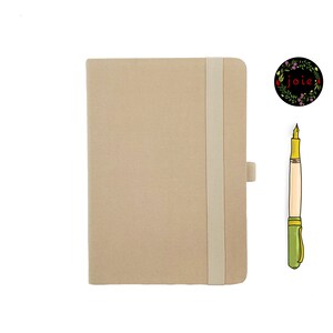 May include: A beige notebook with a white elastic closure and a pen with a green and gold design. The notebook has the word "joie" written in a floral wreath.