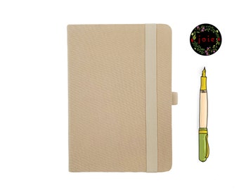 May include: A beige notebook with a white elastic closure and a pen with a green and gold design. The notebook has the word "joie" written in a floral wreath.