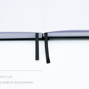 May include: A white notebook with black dotted pages lies flat open. It has two black ribbon bookmarks.