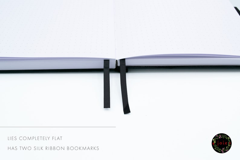 May include: A white notebook with black dotted pages lies flat open. It has two black ribbon bookmarks.