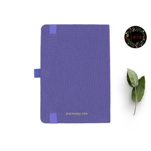 May include: A purple notebook with a gold inscription that reads "joiethings.com". The notebook has a blue elastic band closure and a small blue ribbon bookmark.