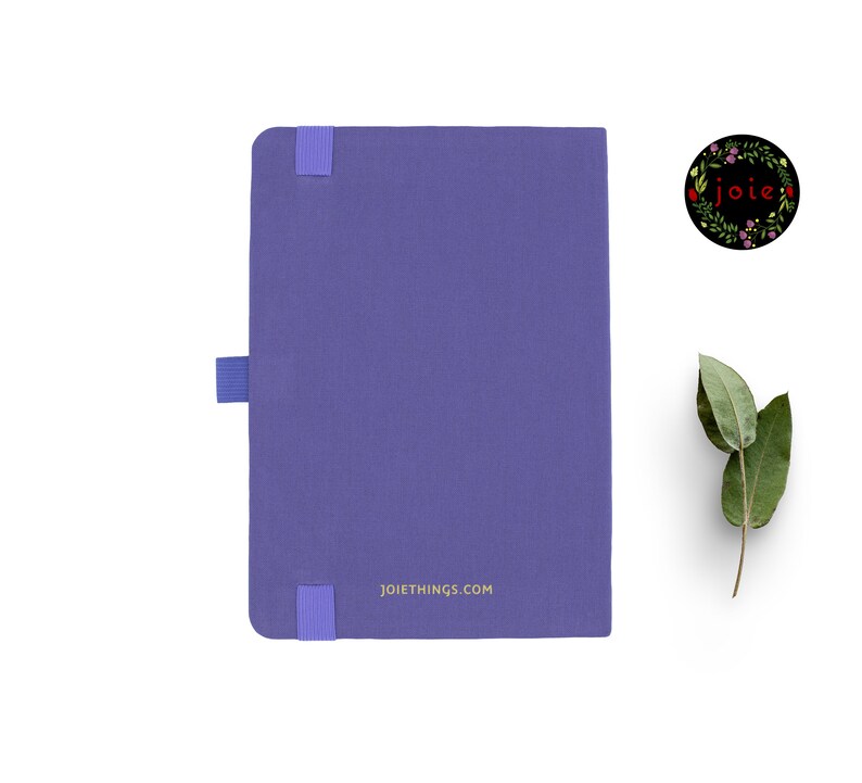 May include: A purple notebook with a gold inscription that reads "joiethings.com". The notebook has a blue elastic band closure and a small blue ribbon bookmark.