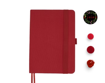 Puede incluir: Un cuaderno rojo con cierre elástico negro y un marcapáginas de cinta roja. El cuaderno tiene una portada lisa y la palabra "joie" está escrita en una corona de flores sobre un círculo negro.