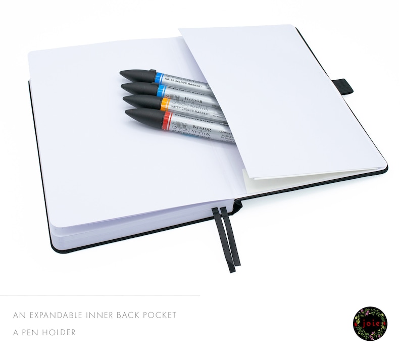 May include: A black notebook with an expandable inner back pocket and a pen holder. The notebook is open and has a white interior. There are four markers in the pocket, including a blue, orange, and two grey markers. The markers have the text "Winsor Newton Water Colour Markers" printed on them.