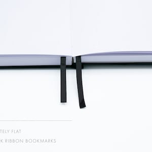 May include: A white notebook with black dotted pages lies flat open. It has two black ribbon bookmarks.