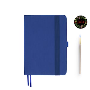 May include: A blue notebook with a black elastic closure and a blue ribbon bookmark. The notebook has the word "joie" written in red on a black circle with floral accents.