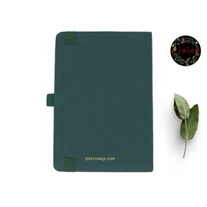 May include: A dark green notebook with a black elastic band closure and a gold inscription that reads "JOIETTHINGS.COM".