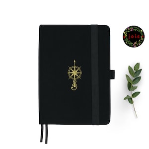 May include: Black notebook with a gold compass design and a black elastic closure. The notebook has a black ribbon bookmark.