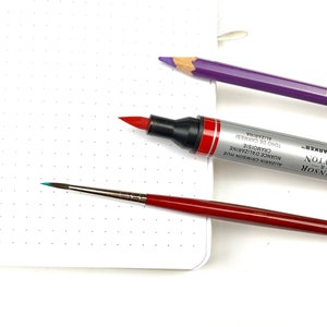 May include: A close-up of three art supplies: a red marker labelled "Winsor & Newton Water Colour Marker", a purple pencil labelled "Hoon-1-Hon", and a small red and silver paintbrush with a turquoise tip.