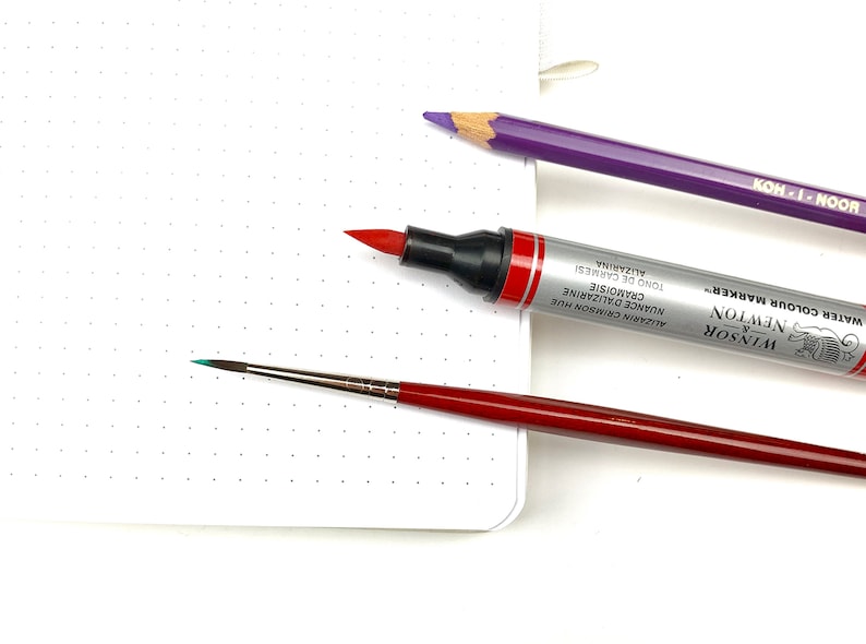May include: A close-up of three art supplies: a red marker labelled "Winsor & Newton Water Colour Marker", a purple pencil labelled "Hoon-1-Hon", and a small red and silver paintbrush with a turquoise tip.