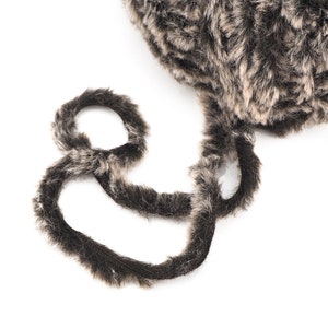 May include: Close-up of a brown and gray faux fur yarn with a long, fluffy texture. The yarn is coiled into a ball and a strand is pulled out, showing the soft, fuzzy fibers.
