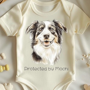 Könnte beinhalten: Cremefarbener Baby-Strampler mit einem lächelnden Porträt eines Australian Shepherd-Hundes. Der Text "Protected by Mochi" ist unter dem Hunde-Bild aufgedruckt. Der Strampler ist von Spielzeug und Dekorationselementen umgeben.