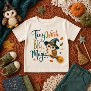 May include: A cream-colored short-sleeve t-shirt featuring a graphic of an owl wearing a witch hat and riding a broomstick. The text reads Tiny Witch, BIG Magic. The design includes small colorful stars and autumn leaves in orange and green.
