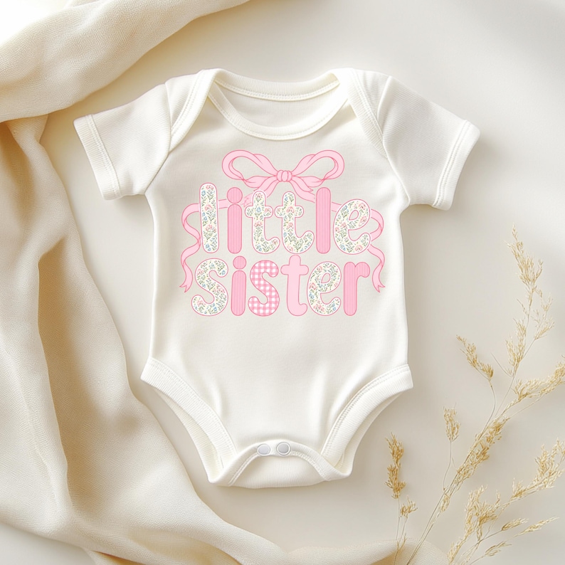 May include: White baby onesie with the words "little sister" in pink floral and gingham print. A pink bow and ribbon accent the text. The onesie has short sleeves and snap closures.