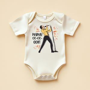 Puede incluir: Body de bebé color crema con un gráfico de un cantante con chaqueta amarilla y pantalón negro, sosteniendo un micrófono. El texto "MAMA! OO-OO-OOH!" está impreso en negro. El diseño está rodeado de detalles en naranja y rojo.