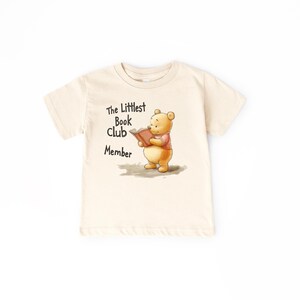 May include: Cream-colored t-shirt featuring a cartoon illustration of a bear reading a book. The text "The Littlest Book Club Member" is printed above the bear. The bear is wearing a pink shirt.