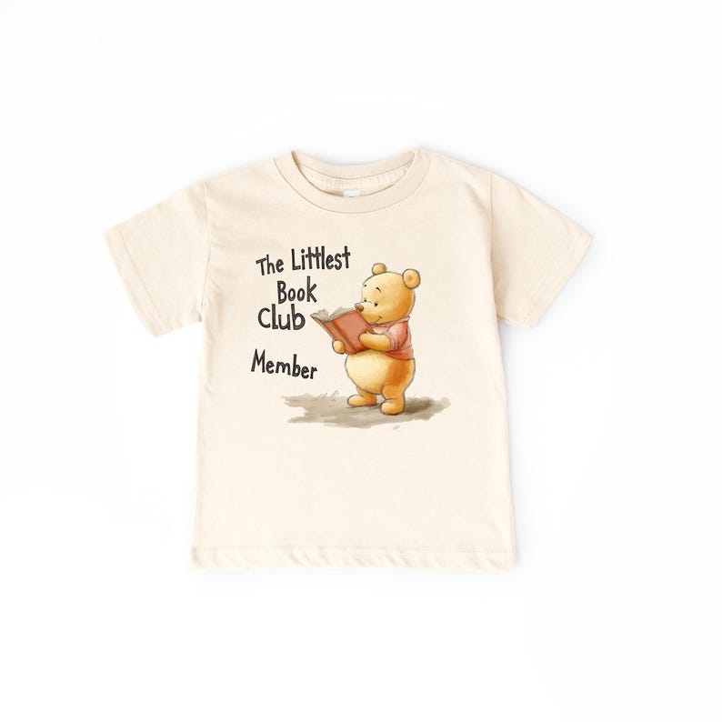 May include: Cream-colored t-shirt featuring a cartoon illustration of a bear reading a book. The text "The Littlest Book Club Member" is printed above the bear. The bear is wearing a pink shirt.