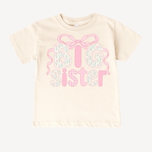 May include: Cream-colored t-shirt featuring the words "BIG SISTER" in pink. The letters "B" and "G" are filled with a floral pattern, while the "I" is a solid pink stripe. The "SISTER" is in pink with a gingham pattern for the "S" and floral pattern for the "I", "S", "T", "E", and "R". A pink bow and ribbons accent the top.