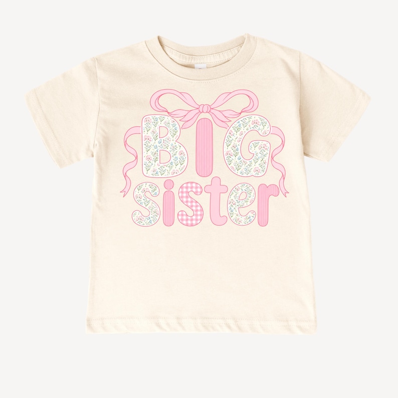 May include: Cream-colored t-shirt featuring the words "BIG SISTER" in pink. The letters "B" and "G" are filled with a floral pattern, while the "I" is a solid pink stripe. The "SISTER" is in pink with a gingham pattern for the "S" and floral pattern for the "I", "S", "T", "E", and "R". A pink bow and ribbons accent the top.